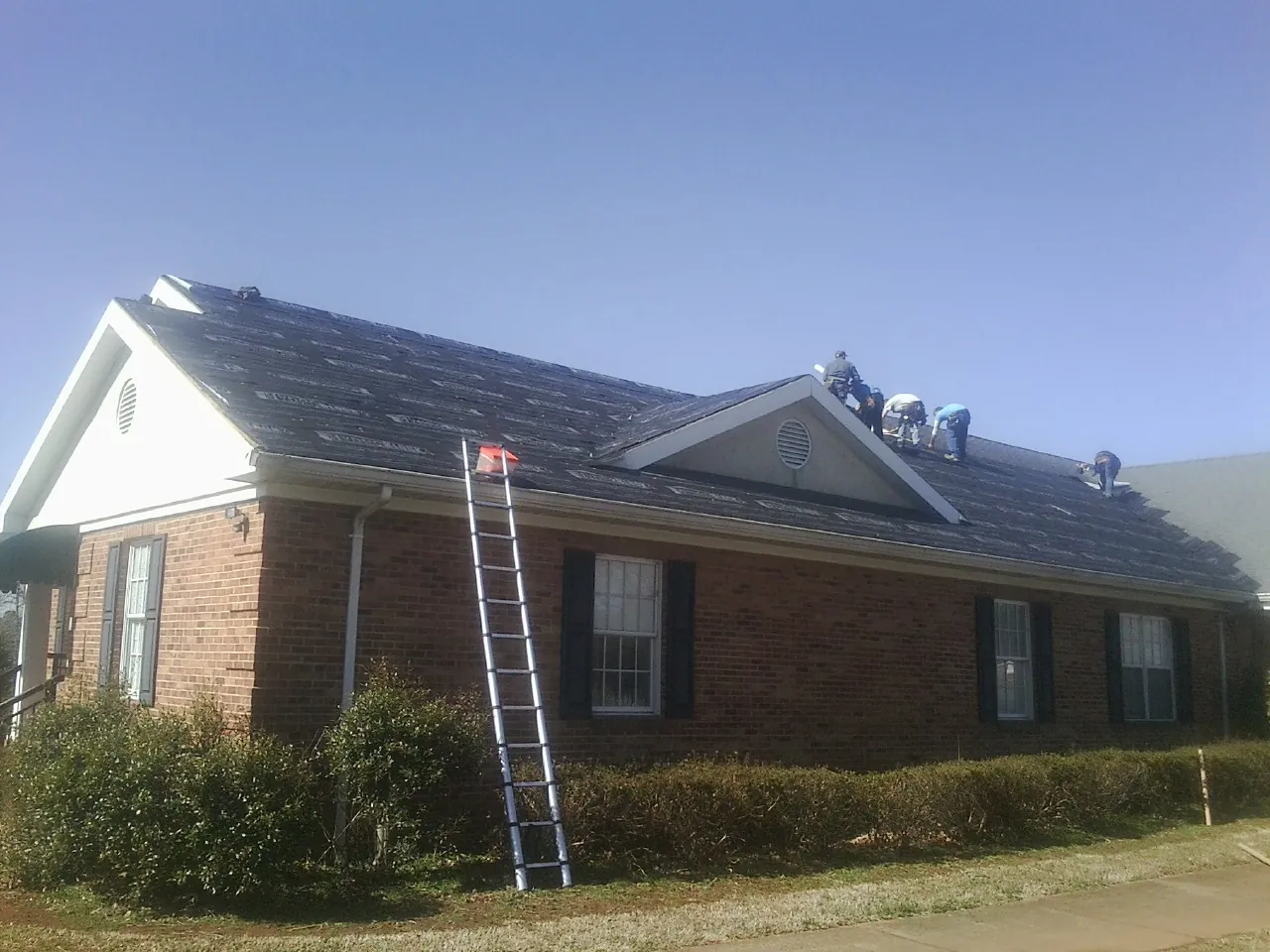 Crew tearing shingles off a brick ranch roof with a ladder set — Metal Roof Repair work by our crew in Tecumseh